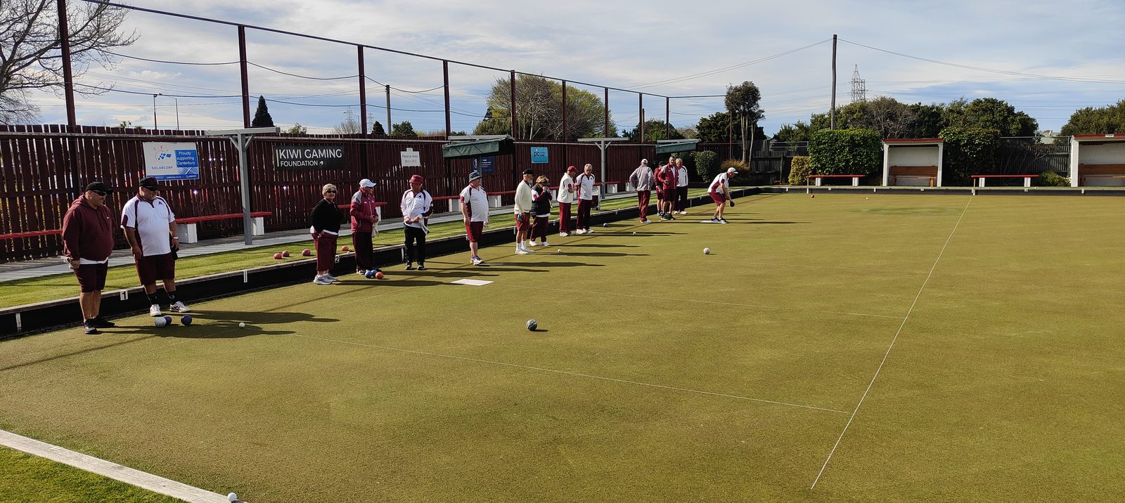 Members of Bowls Hornby in club colours lined up along the rinks, one bowler delivering.